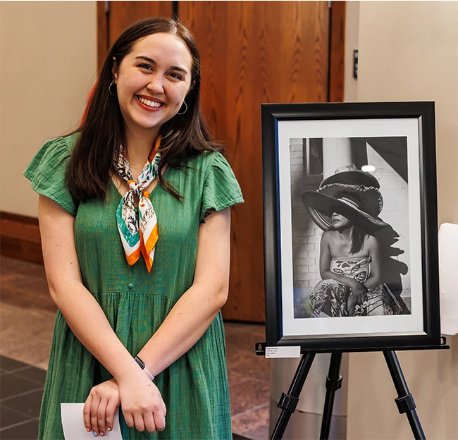 A female student stands beside a framed black and white photograph of a women in a large wide brimmed hat