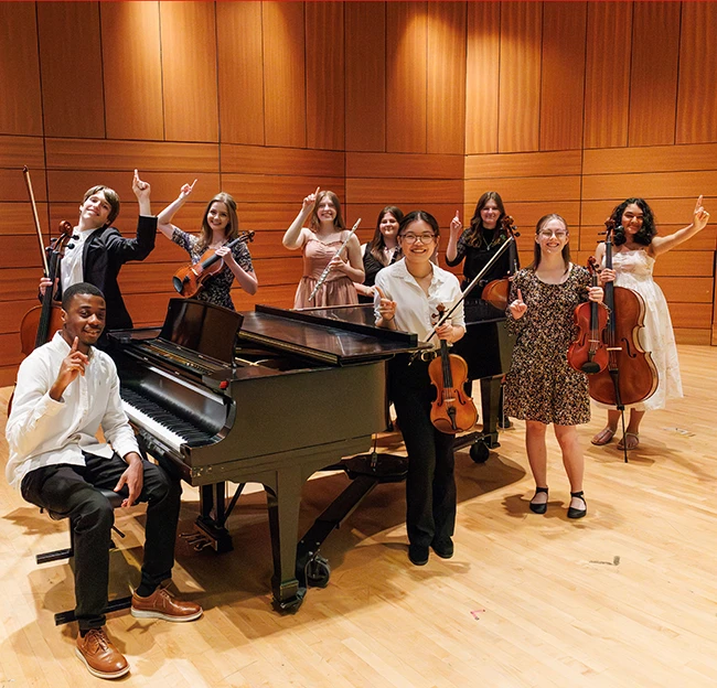 Students gather around a piano on stage holding their instruments with their index fingers raised to indicate number 1