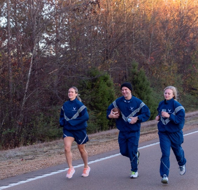 three cadets jogging down the road with one carrying a football