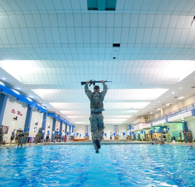 cadet in uniform jumping into pool while holding rifle above head