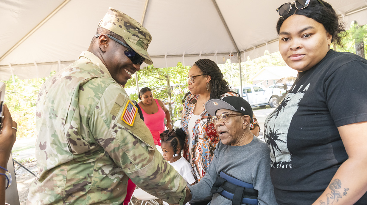 A man wearing Army fatigues shakes hands with a man sitting in a wheelchair uunderneath a pop-up tent.