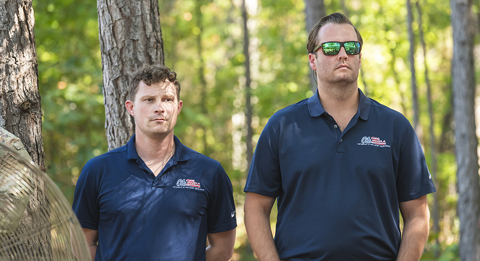 Two men wearing matching blue shirts and sunglasses watch an outdoor ceremony.