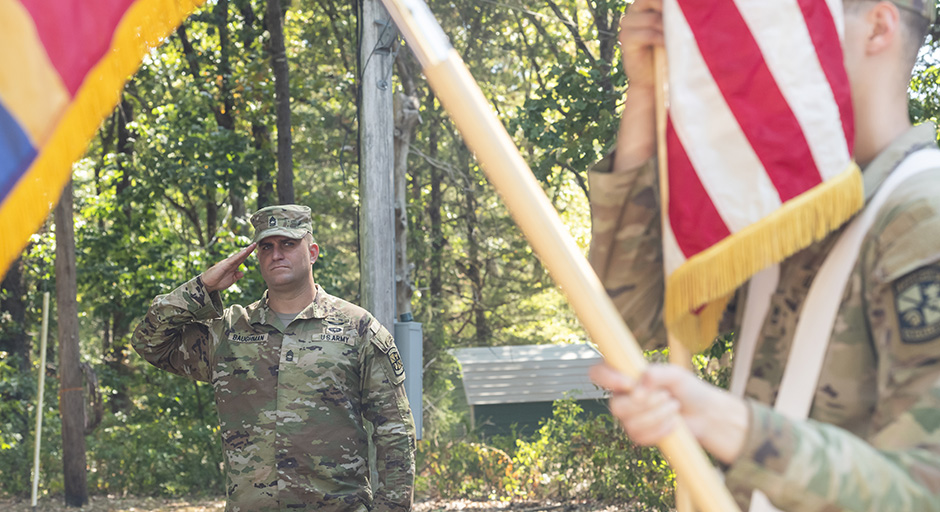 A man wearing Army fatigues salutes as cadets present the American flag at an outdoor ceremony.