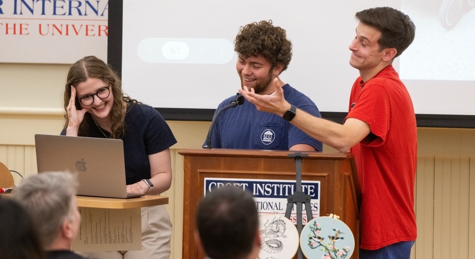 three students standing in front of a crowd. they are smiling and having fun while making a presentation