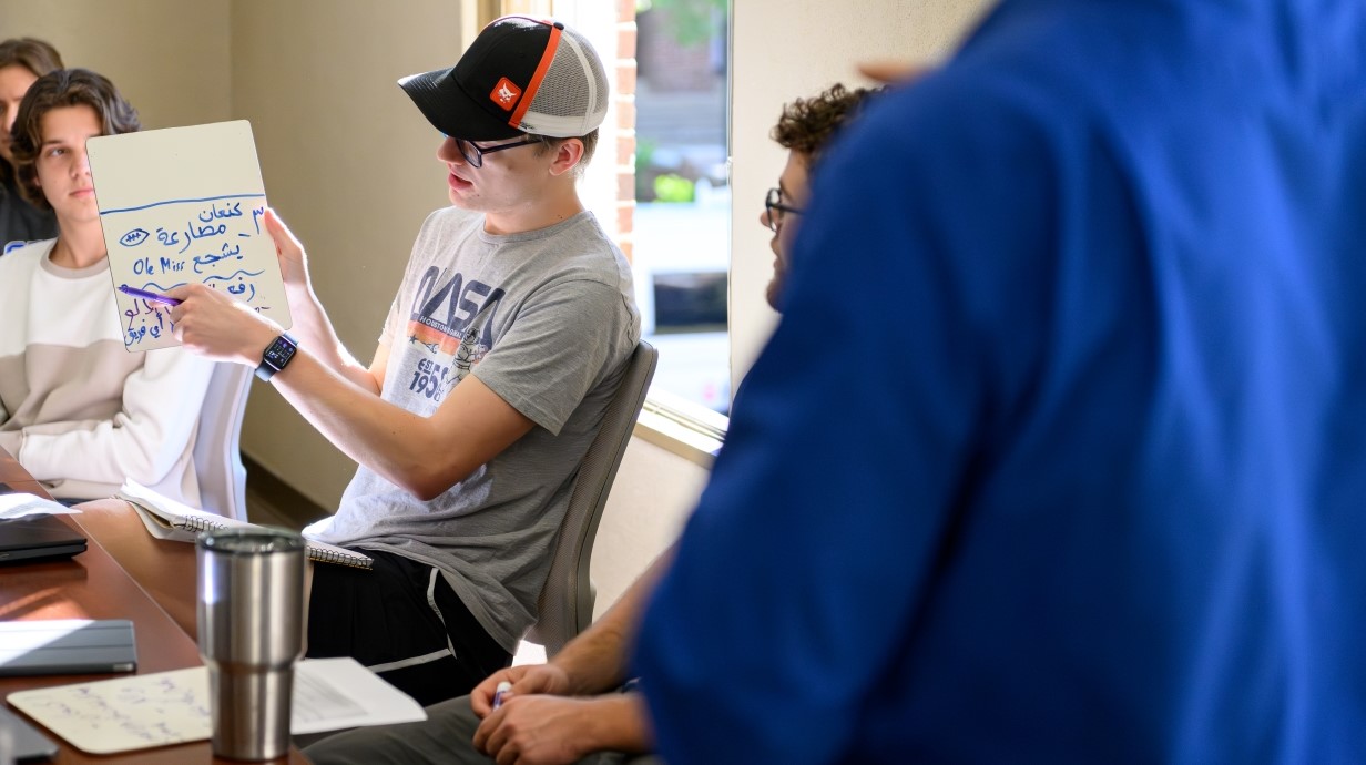 image of a student holding up a small whiteboard in a classroom and pointing to arabic characters.