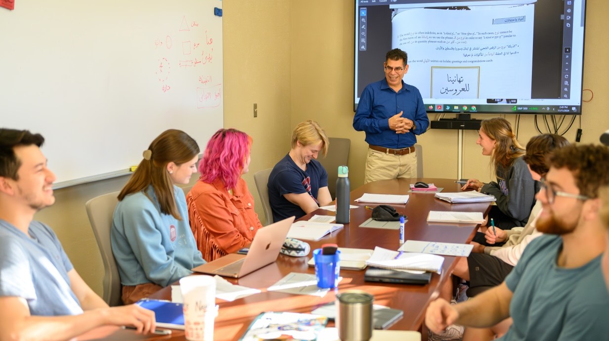 students seated around a table in a class with a professor standing at the end of the conference table