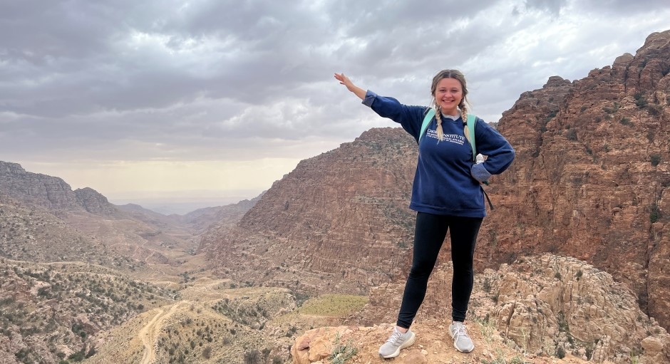 student standing on a mountain ledge with a wide view of dry mountain valley behind her