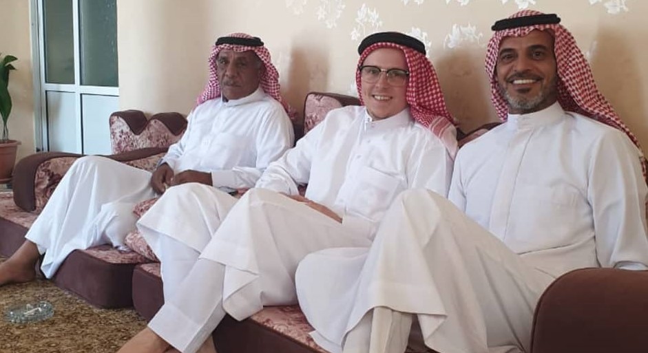 student seated on a couch with two men. all are dressed in traditional clothing and head dress for the Middle Eastern culture