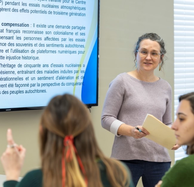 professor standing in front of the whiteboard in a classroom along with two students in the foreground of the photo