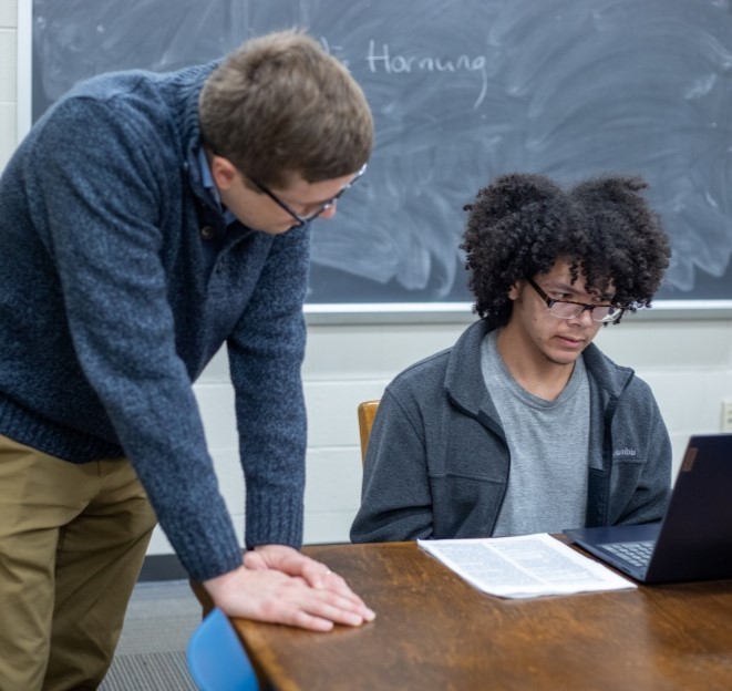 faculty member standing and helping a student seated at the table and looking at a paper together in a classroom