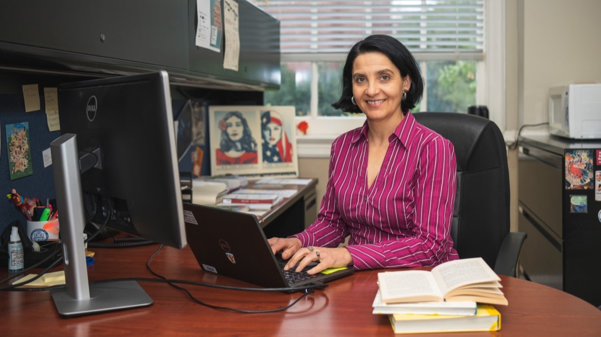 image of professor seated at her desk and smiling at the camera while she sits at her computer