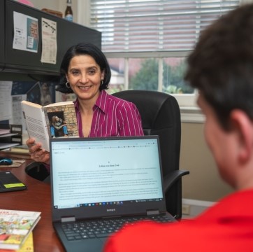 professor seated at a desk and smiling at a student while holding a book