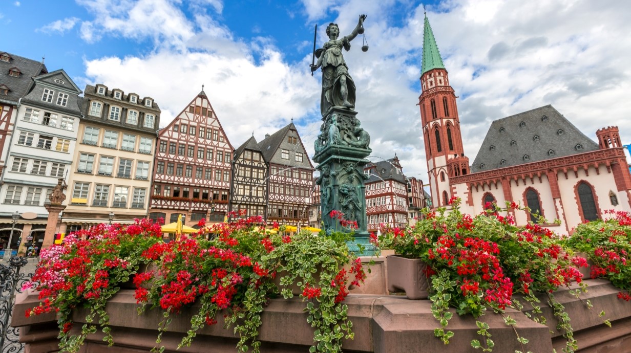 image of a plaza with a fountain, statue of a woman holding the scales of justice, flowers, and historic German buildings in the background