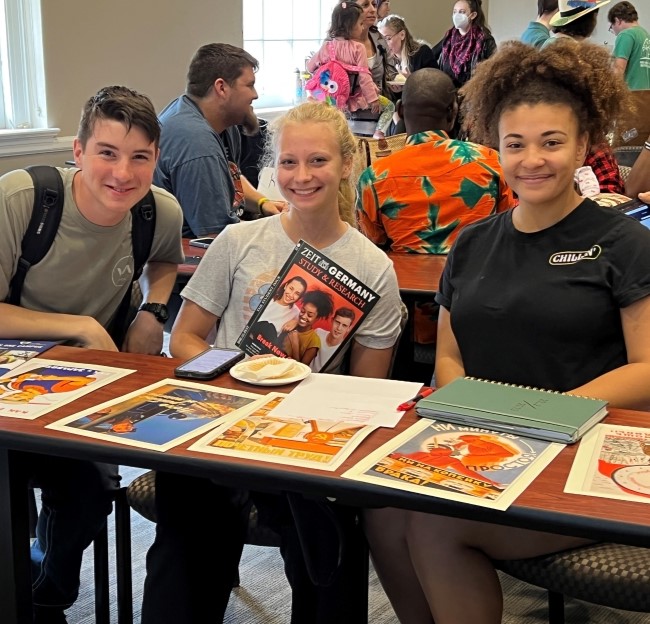 three students seated at a table with German-related papers in front of them on the table