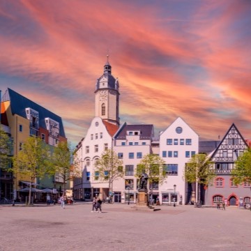 street view of a historic square in Jena with colorful buildings and trees shading sidewalk cafes and a red sunset sky