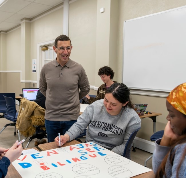 image of a professor standing and smiling at a few students who are seated at a table and using markers to create a poster board