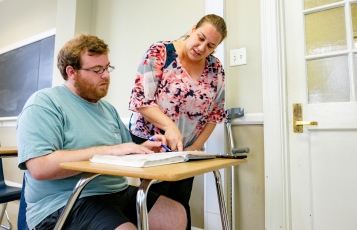 professor standing next to a student seated in a desk; teacher is pointing to the open textbook on the student's desk