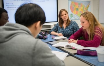 a few students seated around a table with a teacher giving instruction in English