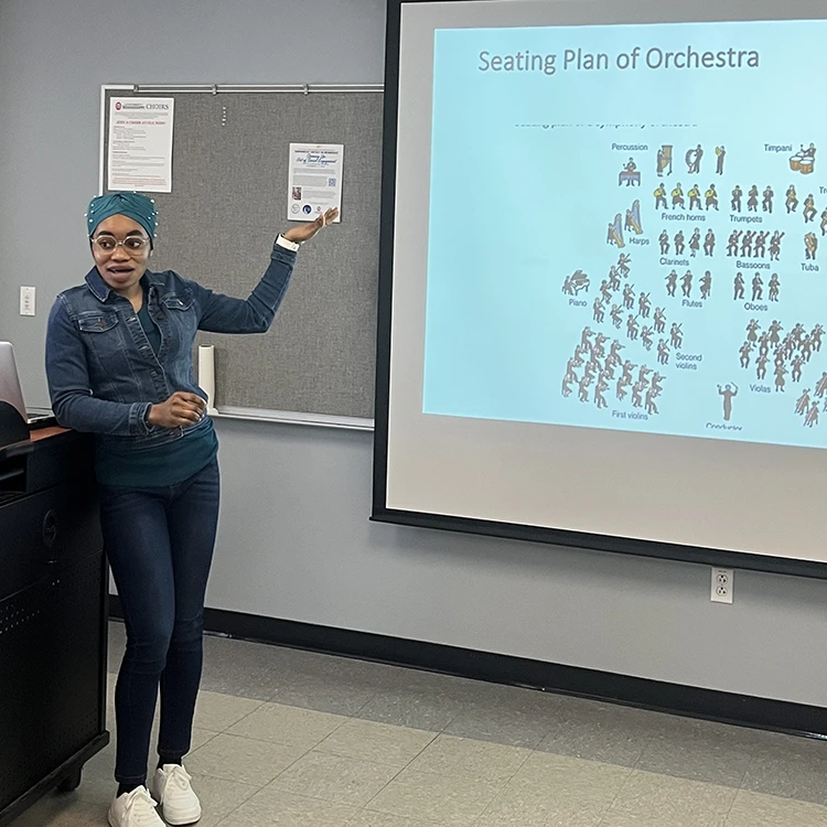 Person leaning on a podium in front of a screen with hand held up. Screen reads "Seating Plan of Orchestra" with diagram of seating 