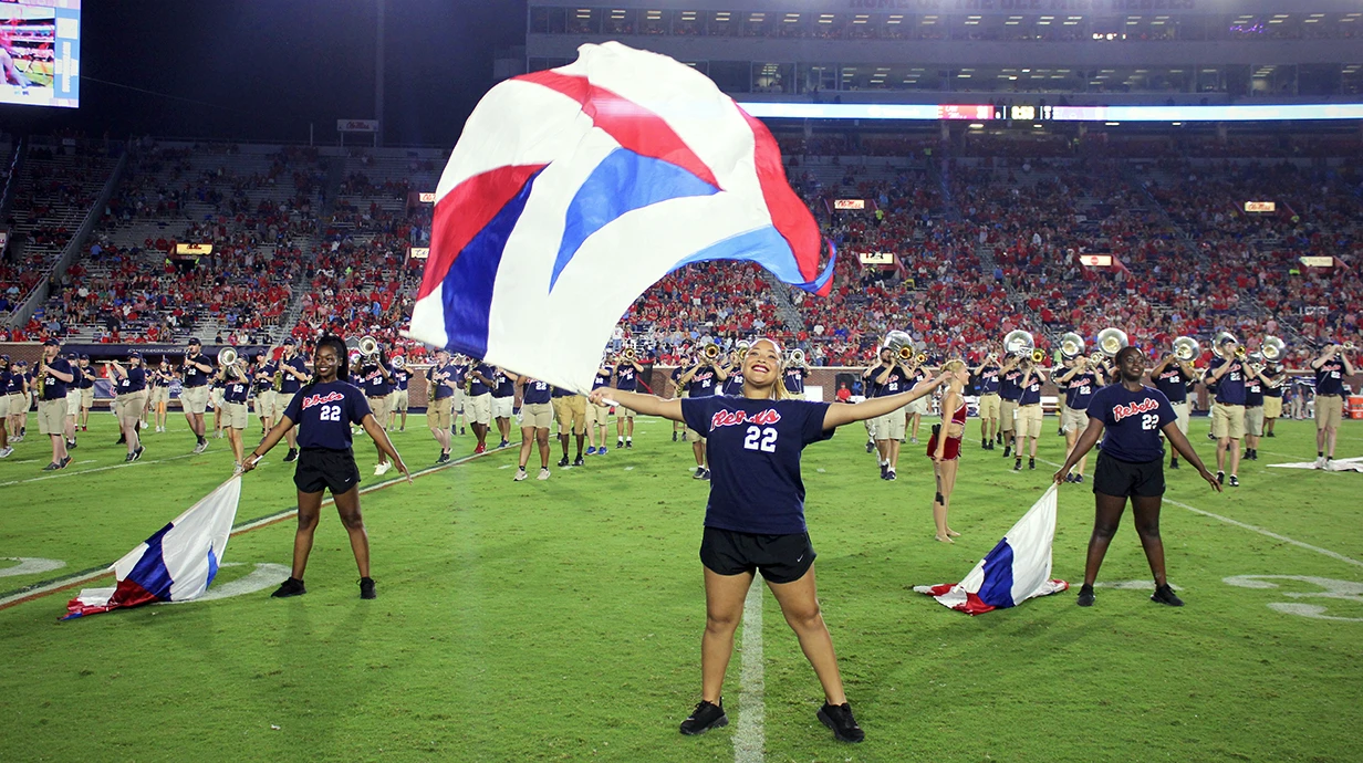 Band members on football field. Camera is focused on person smiling and holding a colorful, unfurled flag