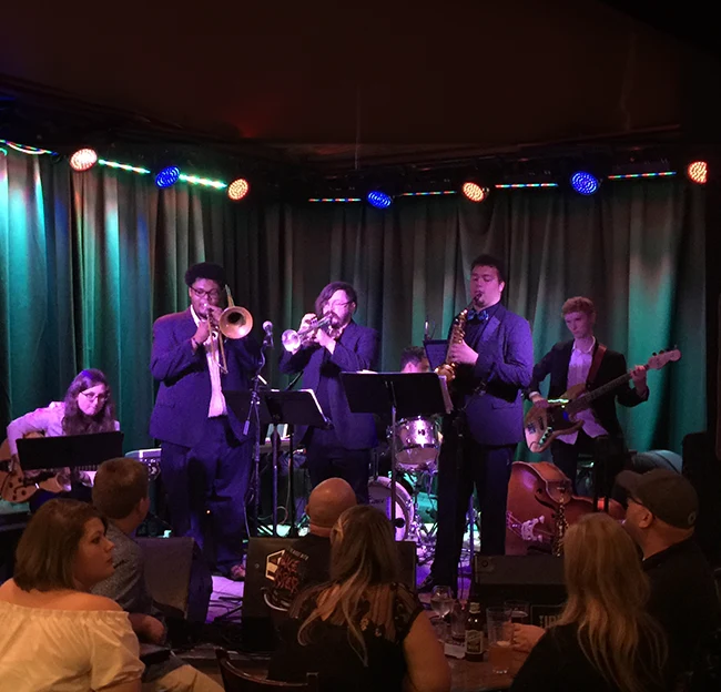 Six piece jazz ensemble perform at darkly lit club. Musicians from left to right play guitar (seated), trombone, trumpet, drums, saxophone, and electric bass guitar.
