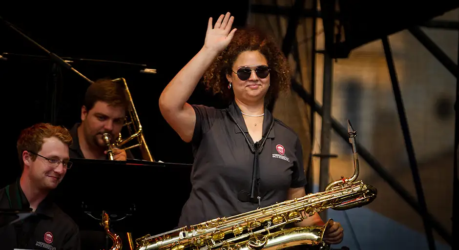Jazz musician on stage with other musicians, wearing sunglasses, holds saxophone and waves at camera.