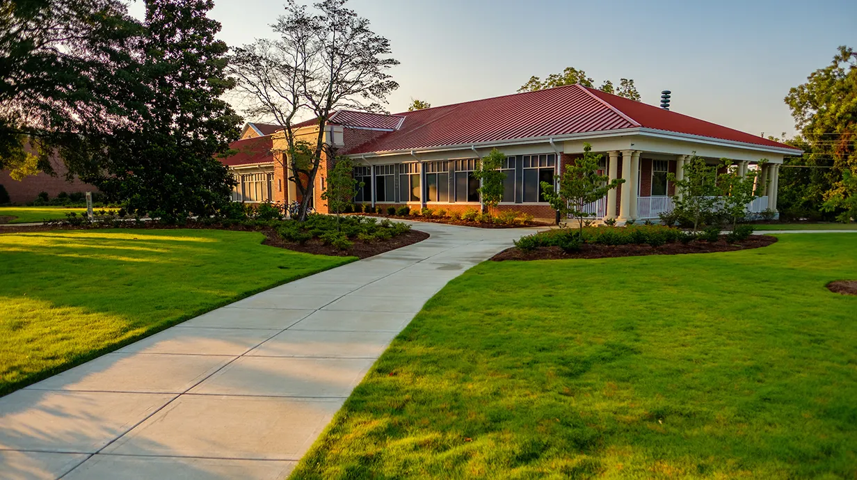 exterior of music building, featuring a red metal roof, green lawn, columns, and a sidewalk.