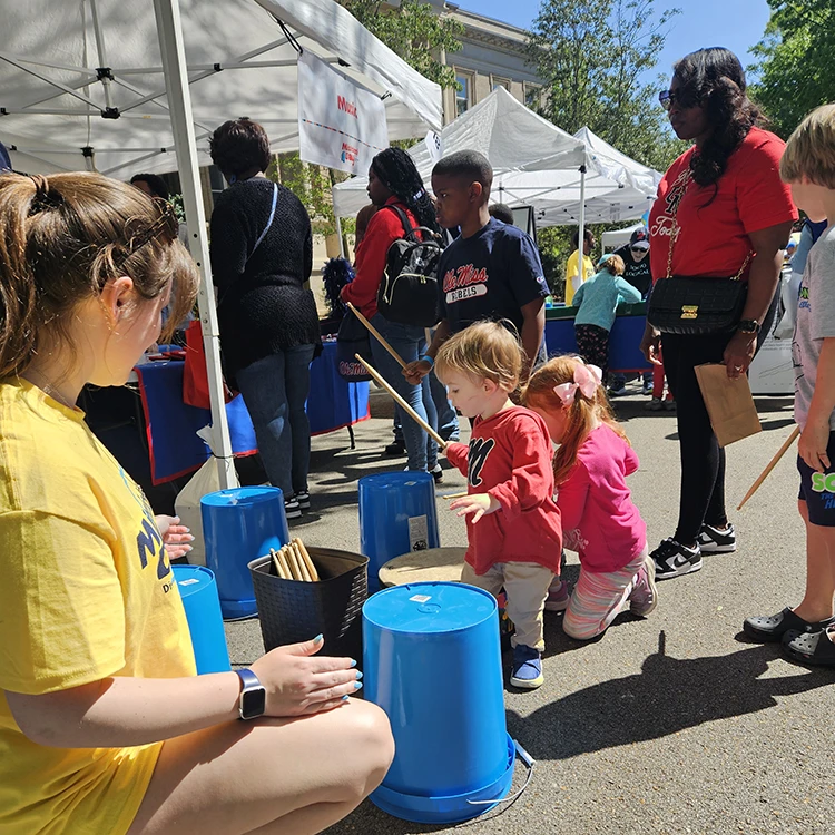 Children and adults in outdoor setting; blue buckets set up as drums