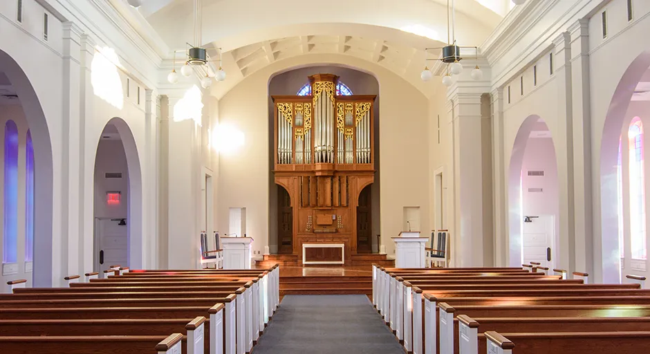 Chapel interior with white vaulted ceilings, wooden pews on either side of an aisle, and floor to ceiling pipe organ in the middle.