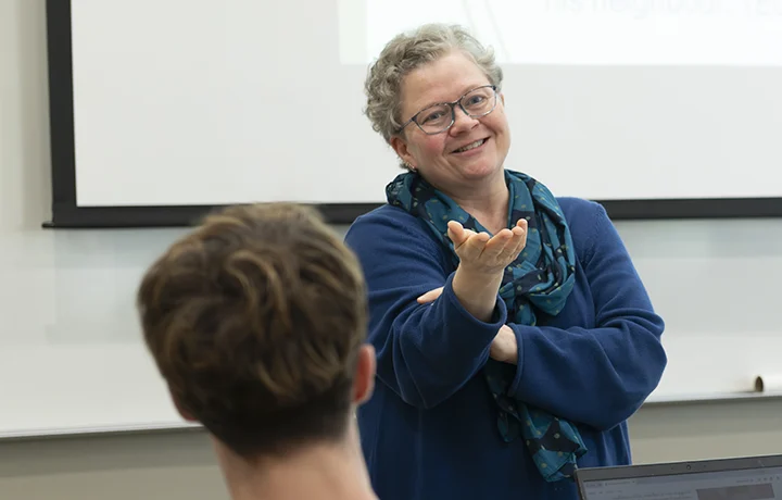 professor stands at front of class, smiling, with hand outstretched