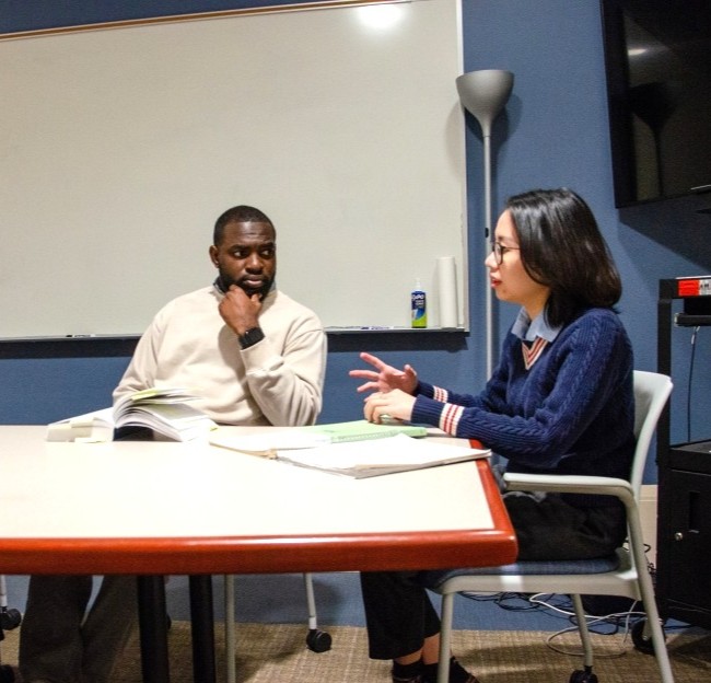 two graduate students seated at a table talking with each other