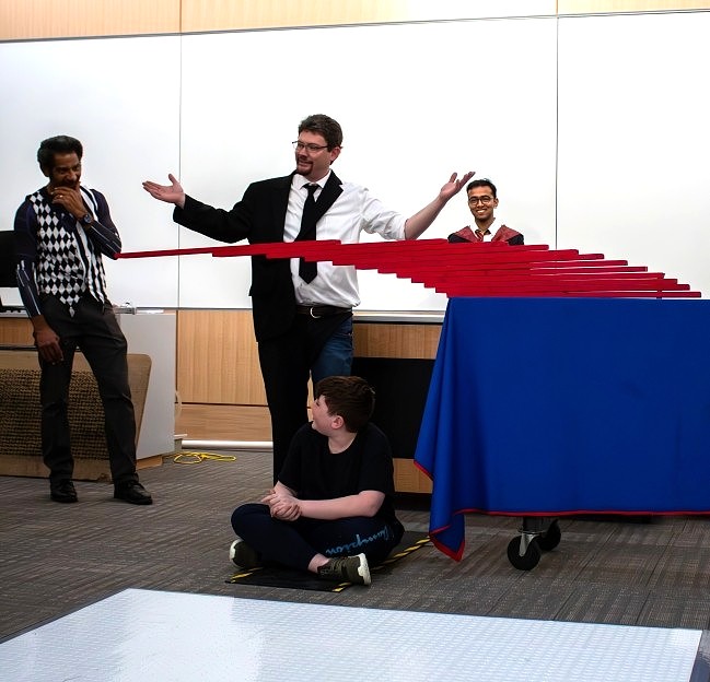 image of a student seated on the floor below a physics demo with planks suspended above him