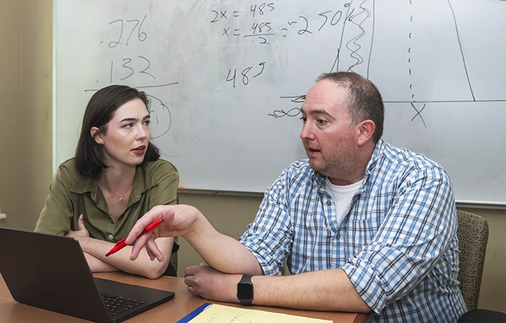 professor and student sit at table looking at a computer; white board with math markings in background