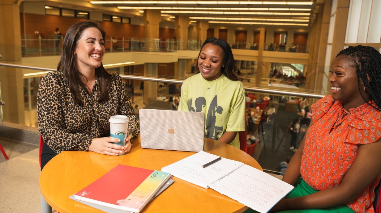 faculty member and two students seated and smiling together in the Union. There is a computer and notebooks in front of them on the table. 