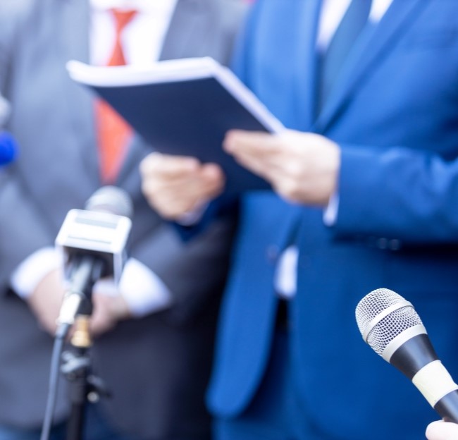 image of reporters holding microphones in front of a man in a business suite who is holding a document as if he is preparing to give a speech.