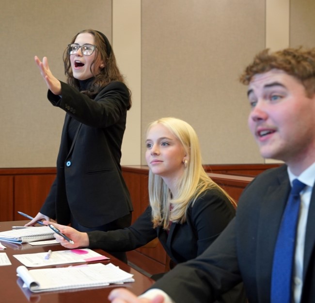 image of Mock Trial Team three members in a courtroom setting gesturing and talking to the witness stand. 