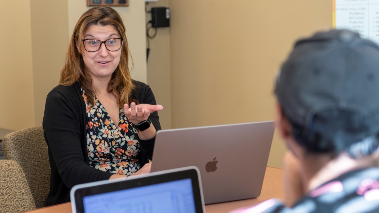 image of professor seated at conference table gesturing and smiling at a student