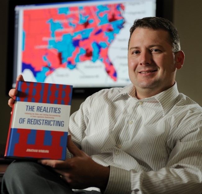 image of Jonathan winburn holding a book for the camera while seated in his office. A political map is on the computer screen behind him