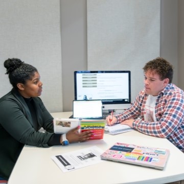 student seated at a table with a writing center mentor with a computer and writing handbook on the table