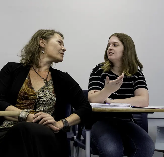 Professor and student sitting in class having conversation. White board in backgroun.