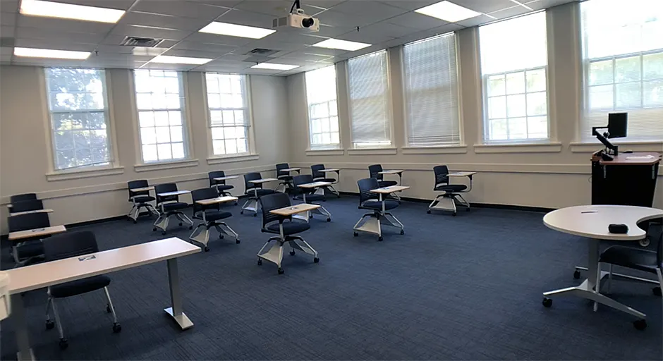 Classroom with fifteen desks for students, blue carpet, a lectern with a computer, and a projector mounted on the ceiling.