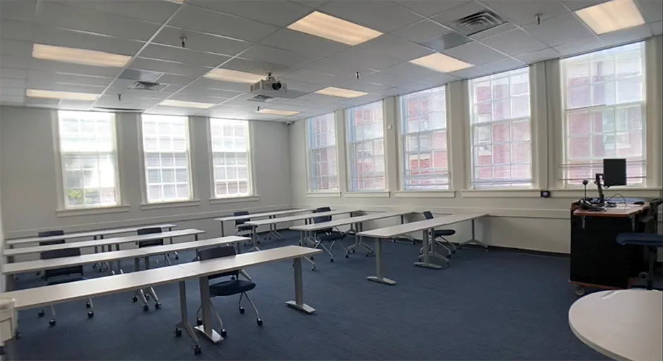 Classroom with long tables and chairs for students, blue carpet, a lectern with a computer, and a projector mounted on the ceiling.