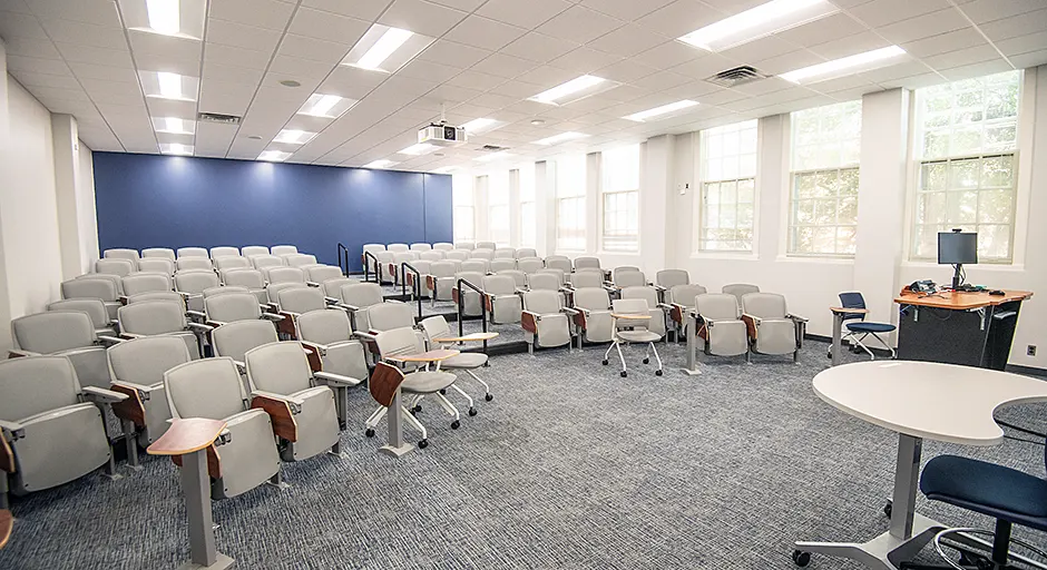 large auditorium with rows of gray seats; a blue accent wall; lectern with computer; and projector mounted on the ceiling.