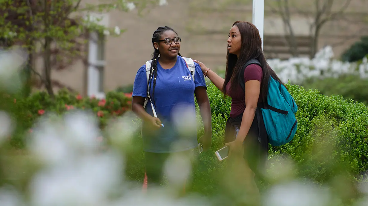 Two students standing outside, brick building in background. One student is talking with their hand resting on the other student's shoulder.