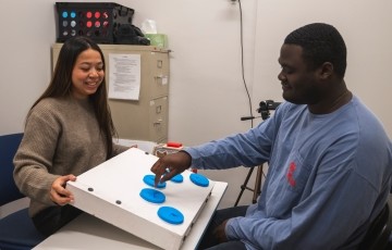 student putting his fingers on a button on a board held up by another student in a research lab. 