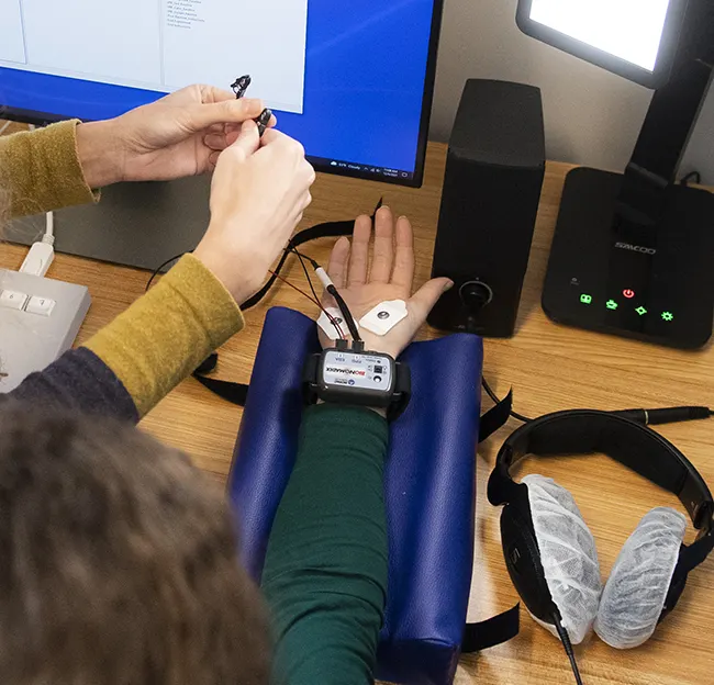 close up of two hands on a desk with wires and electrodes. Computer screen in background and headphones in foreground.