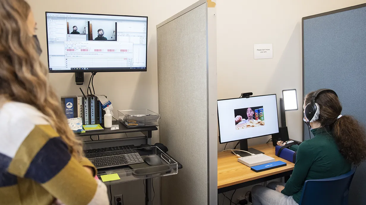 Two people in an office setting. One person is standing behind a monitor and the other person is seated at a computer, hand is connected to a monitoring device, and wearing headphones inside a partition
