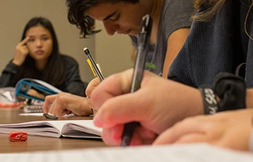 Close up of people studying; hands in foreground holding pens and pencils; writing on paper
