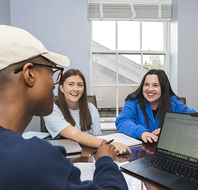three students sitting at a table around a computer smiling