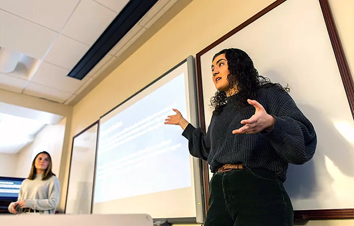 Woman standing at podium speaking and gesturing with her hands. Person on left standing looks at her